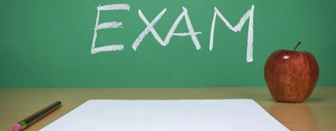 Exam written on a blackboard with an exam sheet, pencil and apple on a table.