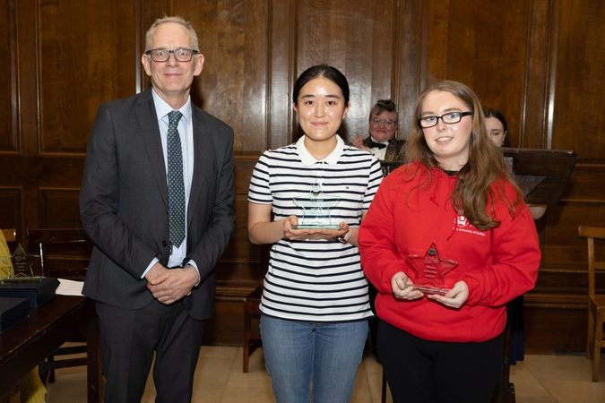 man wearing suit with glasses and tie standing beside two volunteers both holding a star shaped crystal trophy. one is wearing a striped top and the other is wearing a red hoodie