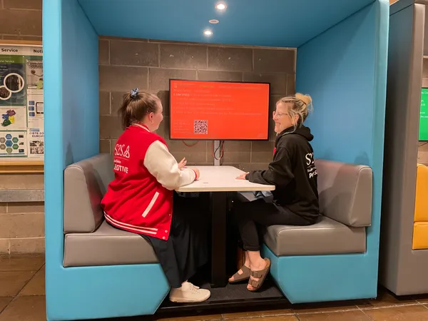 A Head Mentor wearing a red S2S varsity jacket, sits on the left of a square booth. They are chatting with a Peer Supporter who is wearing a black hoodie and sitting on the right.