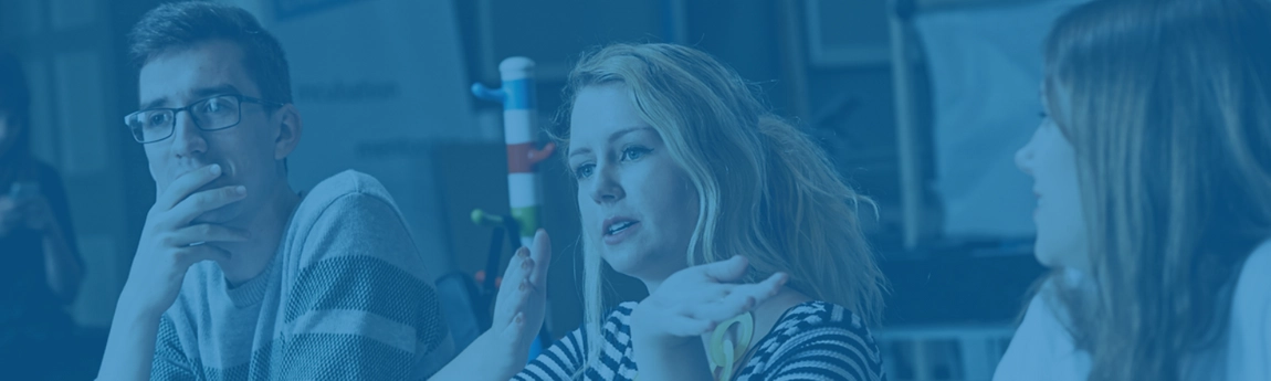 three students sitting at a desk. The middle student is gesturing to something with her hand movement