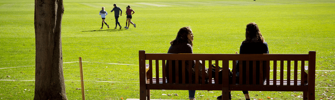 Two people in conversation seated on a bench, on the College sports pitch.