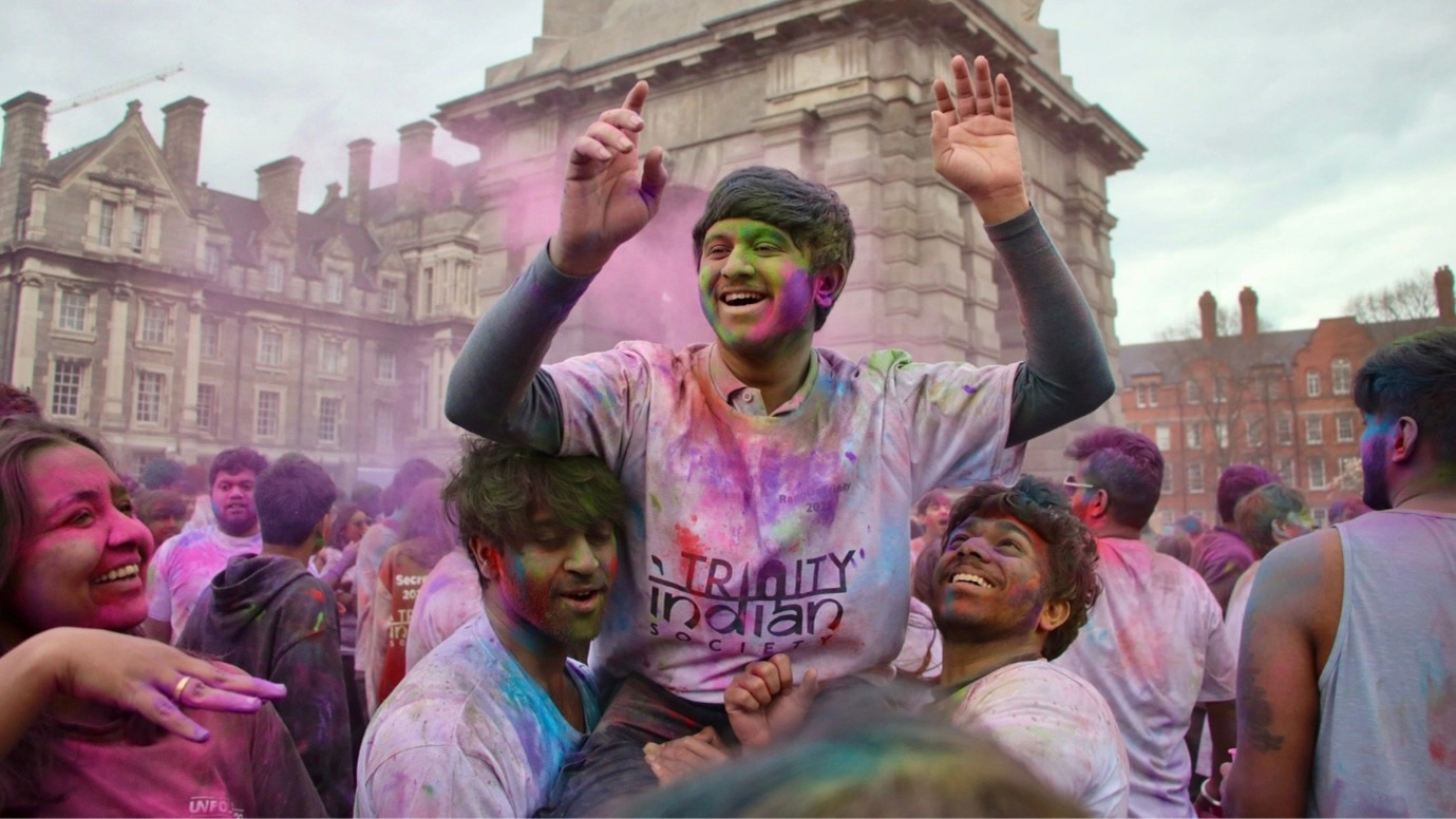 A trio of students from TCD Indian Soc celebrating in a crowd, covered in colourful powder