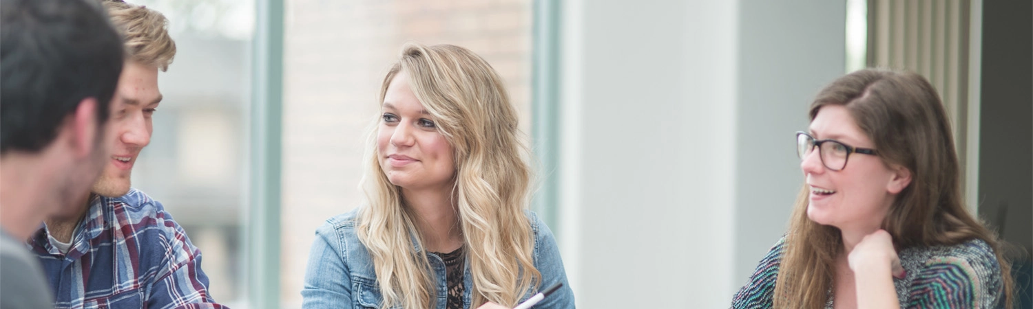 A small group of college students are studying in conference room. They are seated around a table and behind them is a panel of windows facing the outside.
