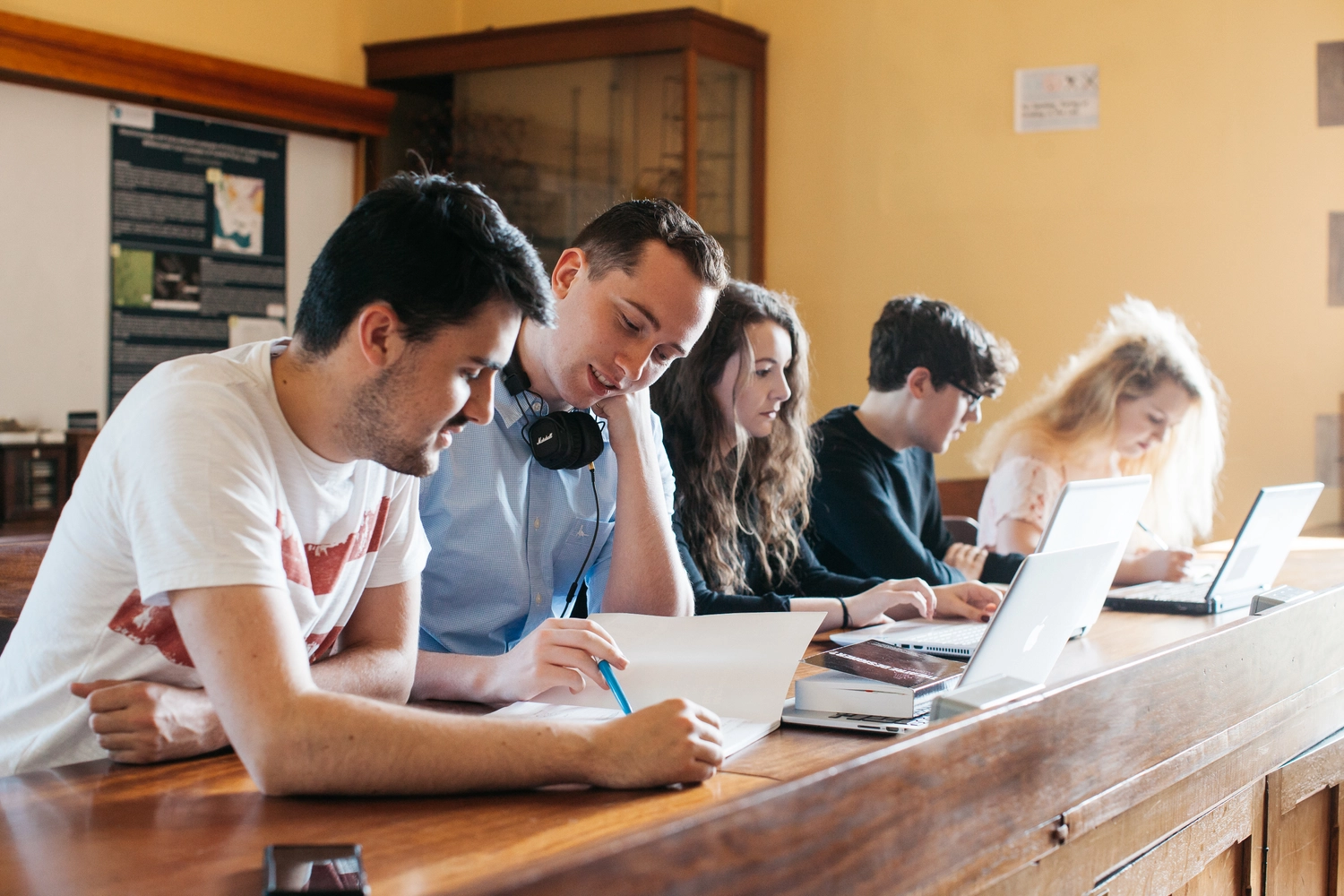 Students sit in a study group together at a long desk using laptops in a bright classroom with large windows and views of trees outside