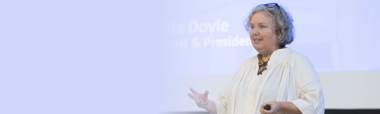 Image of Linda delivering her a speech. She is looking towards the left of the frame at the audience who are not in shot. A screen is partially visible behind her, reading 'Linda Doyle, Provost & President.'