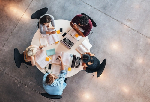 Overhead photo of five students working at a circular table with multiple books open