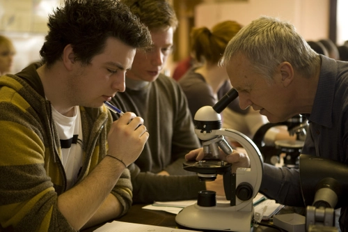 Students at desk in classroom with lecturer looking at microscope and taking notes