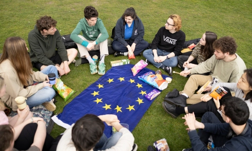A group of students sit in a circle on the grass with snacks placed on a European Union flag in the centre.