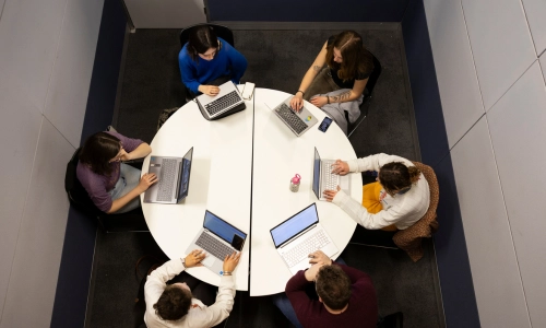 An overhead view of students sitting and talking at a round white table in an open indoor space.