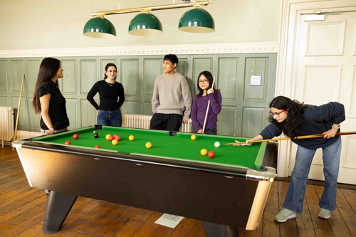 A group of five young adults students gathered around a pool table, with one person aiming to take a shot while others watch and smile