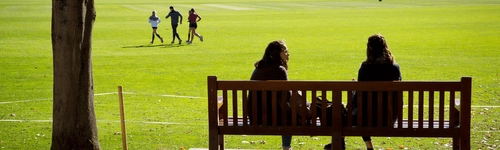 Two people in conversation seated on a bench, on the College sports pitch.