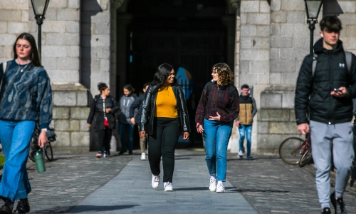 Two students in conversation walking across a paved pathway in Front Square at Trinity College Dublin.