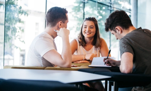 Three students seated together, one student takes notes.