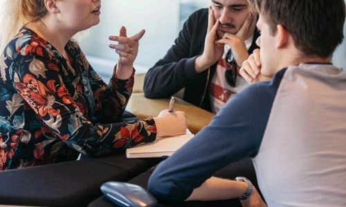 Three students in conversation, one student writes notes.