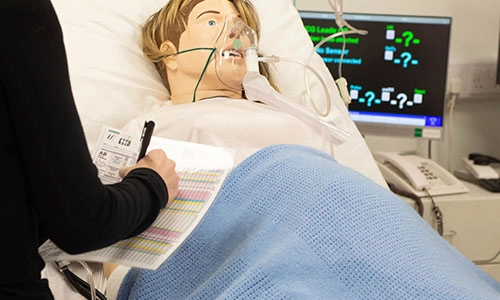 student with a chart beside a medical mannequin in a bed