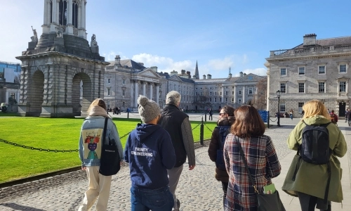Group of people walking through Trinity campus. Their backs are to the camera. Blue skies and Trinity Campanile in the background.