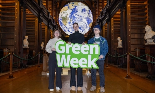 Three students holding a large 'Green Week' sign in Trinity's Old Library. A large globe of the Earth hangs from the ceiling behind them.