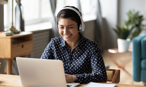 Woman sitting at a laptop with headphones on. She is smiling at the laptop.