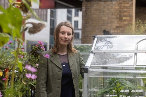 Photo of Eleanor Mullen standing in an urban garden. There is a green house to her right, and pink flowers to her left.
