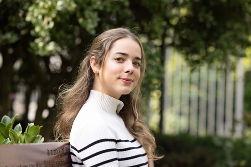 Irene Klotz Maroto sitting on a bench. Photo is taken to her right, and she is looking at the camera. Trees and green foliage blurred in background.