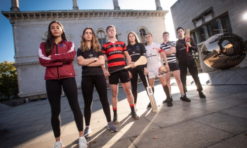 Seven students standing in a line wearing their sport club gear. The student standing in the middle of the row has a hurl in their hand. The last student in the row is holding a cricket bat
