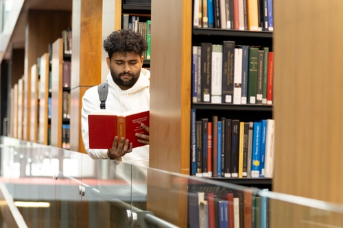 A student stands beside library shelves, holding a red book reading by stacked volumes