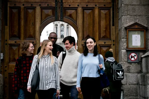 A group of students walk out through large wooden doors of a historic campus building, carrying bags and wearing casual clothing, with the stone archway and detailed windows visible behind them