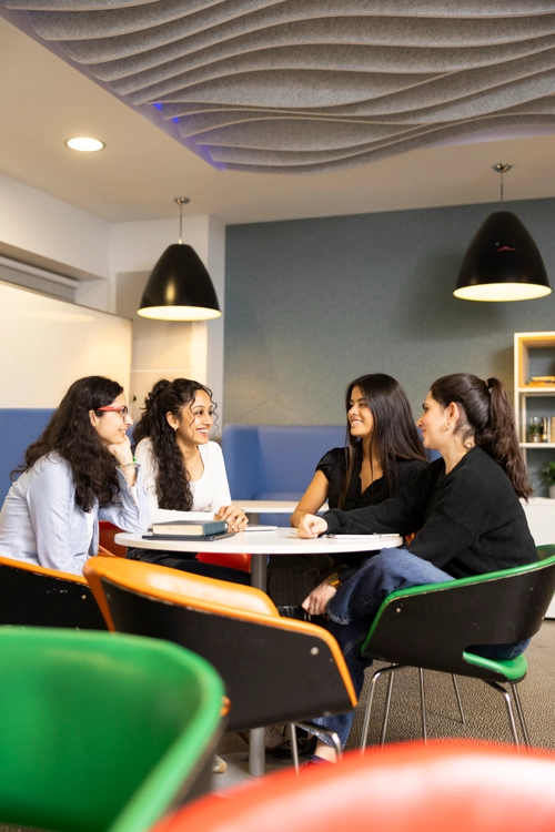 Four students seated around a round table in a collaborative space, having an animated discussion with books and notebooks on the table.