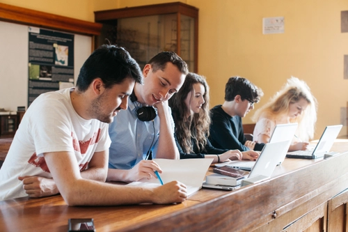 Students sit in a study group together at a long desk using laptops in a bright classroom with large windows and views of trees outside