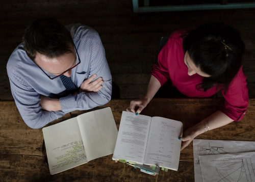 Two students sit at a wooden table reviewing documents, with one open notebook and one open book marked with tab