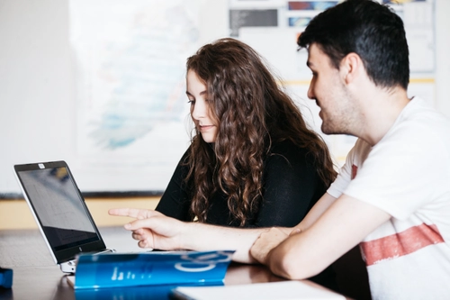 Two students sit together at a desk, engaged while looking at a laptop screen as one points toward it