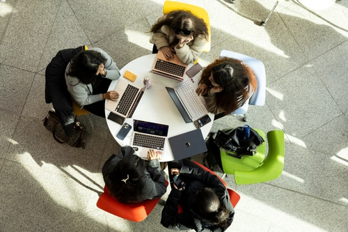 Overhead view of five students sitting around a white circular table, all working on laptops with brightly colored chairs and sunlight on the floor