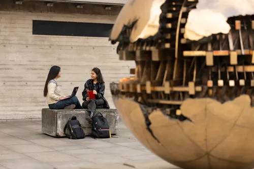 Two students talk while seated on a stone bench with a laptop and notebook, the large bronze Arnaldo Pomodoro sphere sculpture prominently in the foreground.