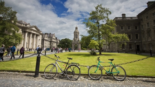 A wide view of Front Square at Trinity College Dublin, showing the Campanile centered in the distance, people walking across the square, and two bicycles chained along the grass in the foreground beneath a partly cloudy sky