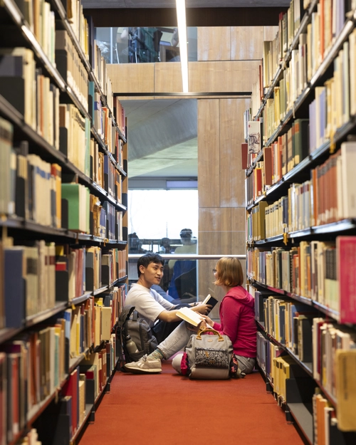 Two students sitting on the floor between tall library bookshelves, studying together with open books