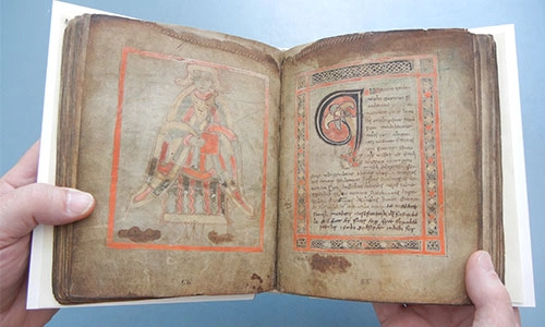 old manuscript with coloured illustrations being opened by pair of hands