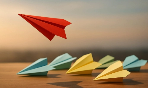 Seven blue, yellow and red paper airplanes lined up on a table, with one red paper airplane in-flight