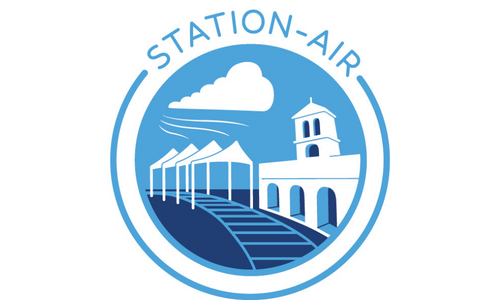 Logo of Station Air. The logo is circular and contains the words station air printed in blue writing with a few images underneath the logo: a cloud with rain dropping from it, a rail track with four marquee-like tents covering the rail track from the rain, and a building with three arches and a bell tower on top