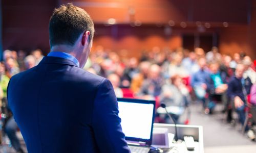 A person speaking to an audience in a lecture theatre. The camera shows the back of the speaker and the audience are blurrred