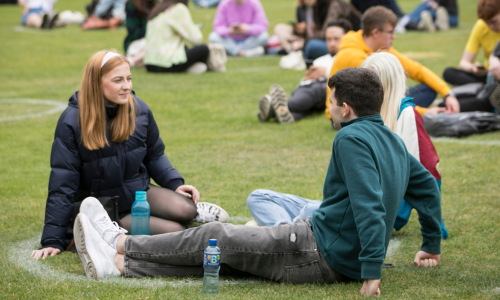 Two Trinity students sitting on grass in conversation.