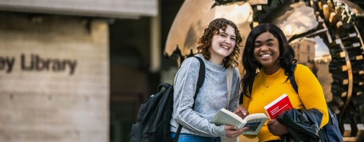 Two students posing holding a book in front of the bronze Globe situated outside the Main Library in Trinity College Dublin