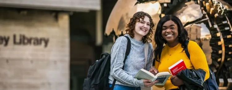 Two students posing holding a book in front of the bronze Globe situated outside the Main Library in Trinity College Dublin