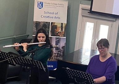 Picture of Vocal Showcase event with acclaimed Irish singer Elizabeth Hilliard and PhD student and flautist Anastasia Motiti performing in front of music stands near a grand piano with a School of Creative Arts banner in the background