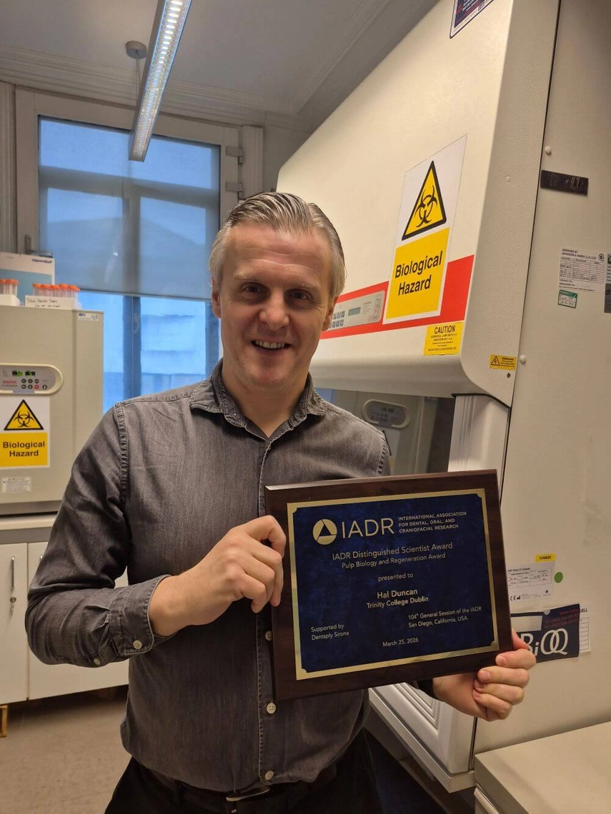 Photo of Prof Hal Duncan holding the IADR award in March 2026. Prof Duncan is standing in a lab in San Diego, California. There are two machines behind him with signs marked Biological Hazard.