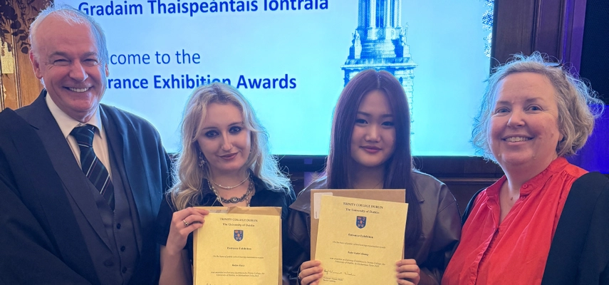 Exhibition Awards in the Trinity Dining Hall, Linda stands for a photo with two students who are holding up their certificates, and Senior Lecturer Vinny Wade. There is a TV screen in the background saying 'Welcome to the Trinity Entrance Exhibition Awards.'