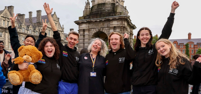 Linda standing with students in front of the Campanile. They are all smiling and cheering for the photo. One student holds Trinity Ted, a large, brown teddy bear.