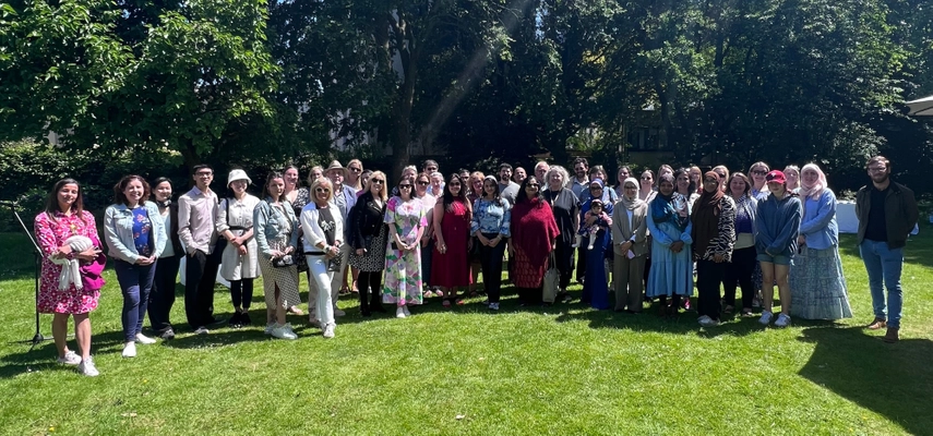 Large group photo taken in a garden. The crowd are standing on grass, with trees in the background and the sun is shining.