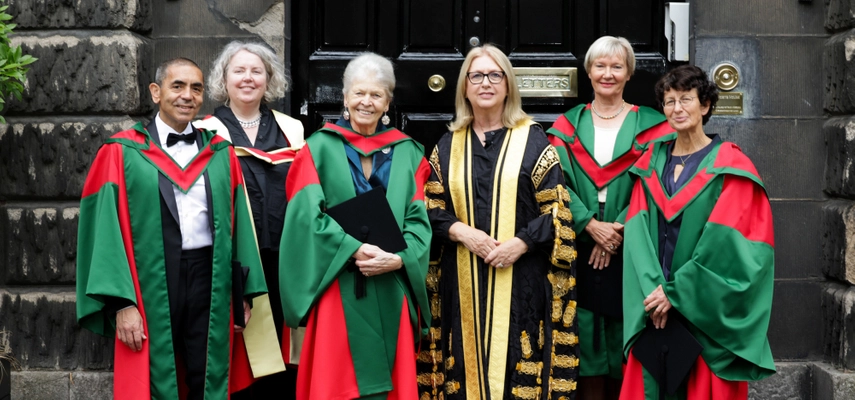Six people standing on stone steps in front of a black door. They are all wearing ceremonial academic gowns.