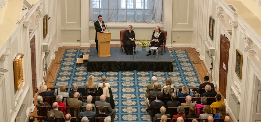 Photo looking down on an event with audience sitting in front of a small stage, with 3 people on it. Two of the people on stage are sitting, and one is standing.