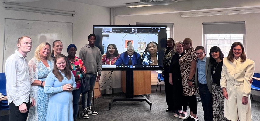 Group of people standing beside a large screen with people on a Zoom call. Everyone is smiling for the camera.